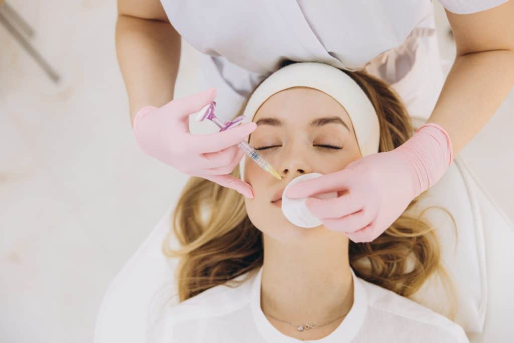 Beautician wearing pink gloves injecting botox filler into woman's lips lying on couch in beauty salon Restylane